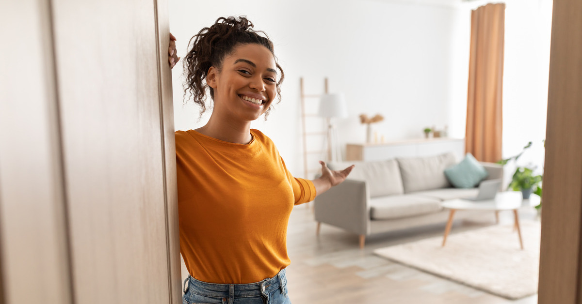 A woman stands behind a door, welcoming the viewer inside. The room behind her has a white couch and coffee table.