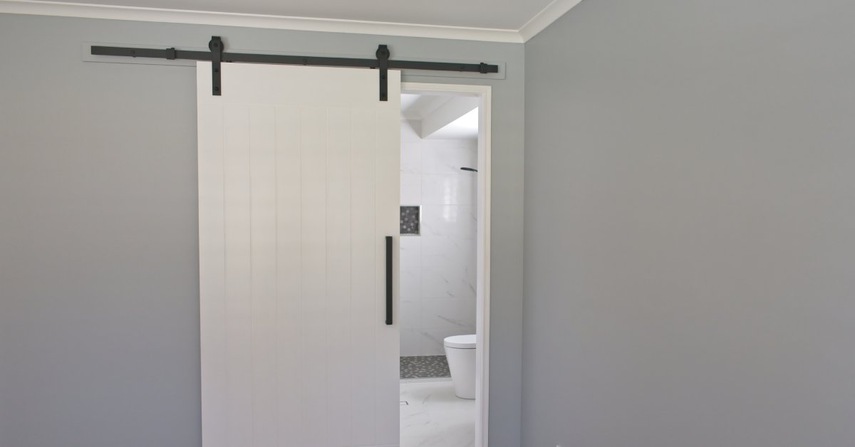 A modern, white, barn style sliding door on a black metal track. Behind the door is a bathroom with a toilet.